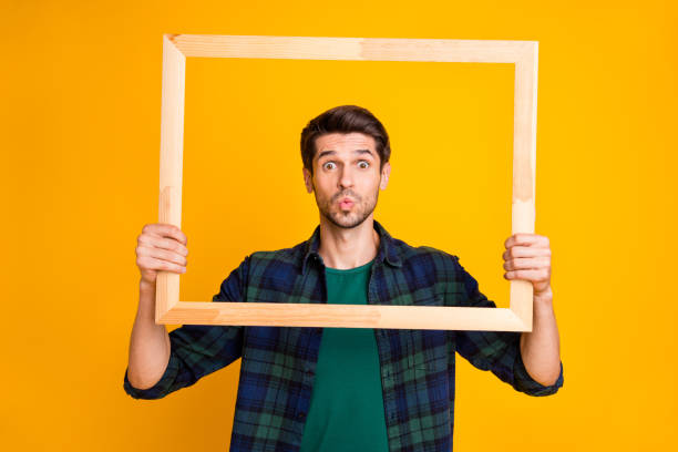 foto de chico divertido sosteniendo marco de imagen de madera mirando a través de ella envío de beso de aire desgaste casual camisa a cuadros de fondo de color amarillo aislado - blowing men kissing blowing a kiss fotografías e imágenes de stock