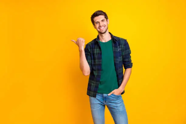 Photo of Photo of amazing guy indicating thumb finger to empty space wear casual plaid shirt and jeans isolated yellow color background