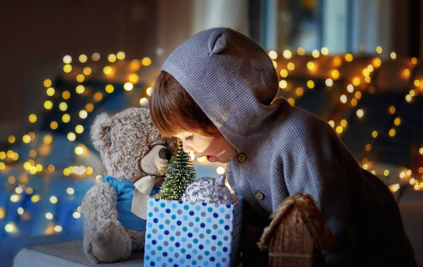 Christmas mood. Cute little excited child with teddy friend looking inside gift box with christmas toys and light from it with garland lights bokeh at background at home. stock photo