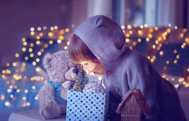 świąteczny nastrój. cute little podekscytowany dziecko z misia patrząc wewnątrz pudełko z zabawkami świątecznymi i światło z niego z girlandą światła bokeh w tle w domu.  fioletowy fioletowy tonowanie magii. - christmas child baby surprise zdjęcia i obrazy z banku zdjęć