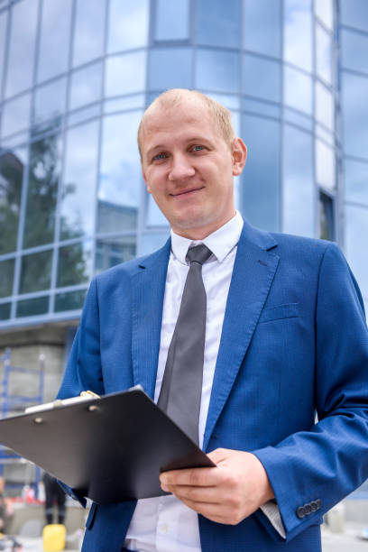 istock Smiling man with clipboard in suit posing before business center 1175674515