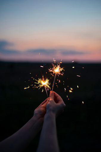 Female hands holding sparkling fireworks to the sky at twilight against sunset sky