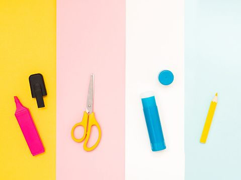 Back to school, education concept. Multicolored school tools; pink marker, yellow scissors; blue glue; white pencils isolated on different colors background with copy space. Flat lay, view from top.