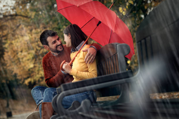 Two young tourists, a man and a woman, are standing on a bridge in london on a rainy day, holding a transparent umbrella and admiring the urban view