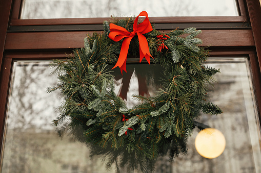 Stylish christmas wreath with red bow and berries at front of store at holiday market in city street. Space for text. Rustic decoration. Christmas street decor.