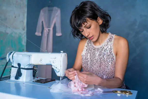 Brunette fashion designer young woman in sewing machine with pink sequins dress at indoor