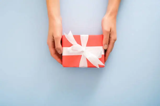 Female's hands holding red gift box with white ribbon bow on pastel blue background. Christmas, New Year, Valentine's day and birthday concept. Minimal flat lay style composition, top view, overhead