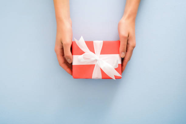 female's hands holding red gift box with white ribbon bow on pastel blue background. christmas, new year, valentine's day and birthday concept. minimal flat lay style composition, top view, overhead - hand wrap imagens e fotografias de stock