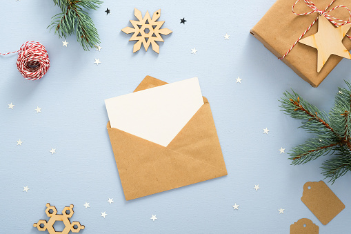 Empty blank card in kraft paper envelope on pastel blue background decorated with confetti star, gift box, clew of rope, fir tree branches. Christmas, New Year, winter holiday invitation mockup.