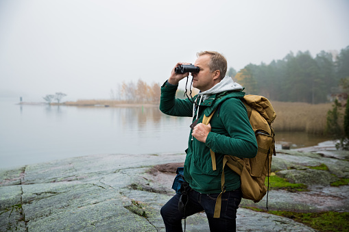 Mature man exploring Finland in the fall, looking into fog through binoculars. Hiker with big backpack standing on mossy rock. Scandinavian landscape with misty sea and autumn forest.