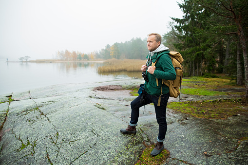 Mature man exploring Finland in the fall, looking into fog. Hiker with big backpack standing on mossy rock. Scandinavian landscape with misty sea and autumn forest.