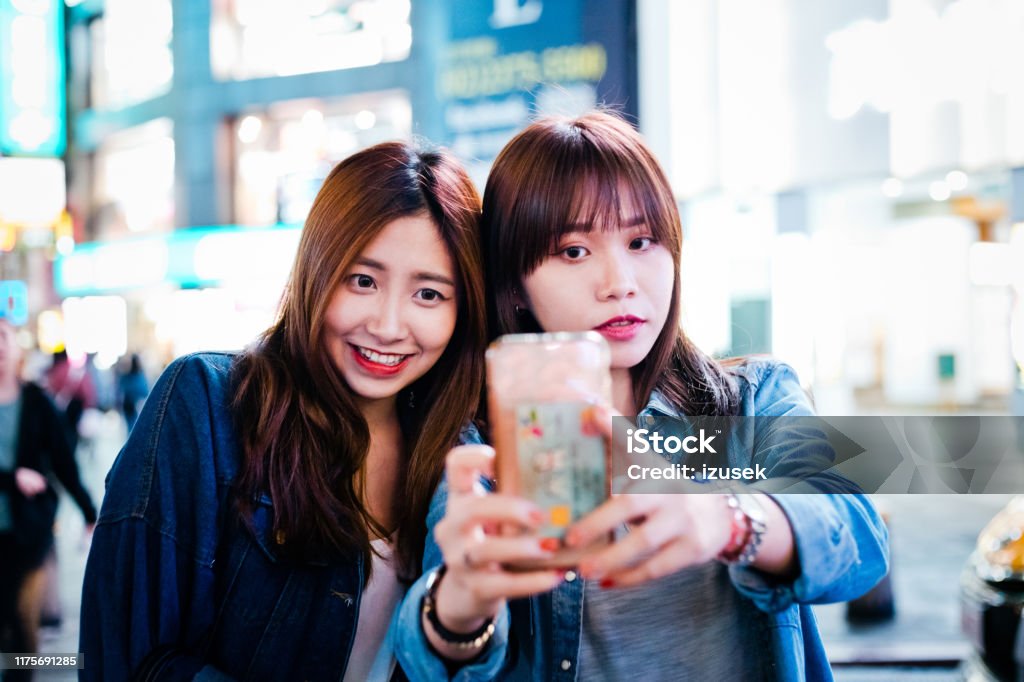 Beautiful female friends taking selfie at night Beautiful female friends taking selfie at night. Young females self photographing in city. They spending weekend together. Asian and Indian Ethnicities Stock Photo