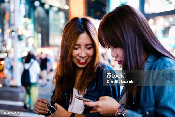 Female Friends Using Smart Phone In City At Night Stock Photo - Download Image Now - 20-24 Years, Adult, Adults Only