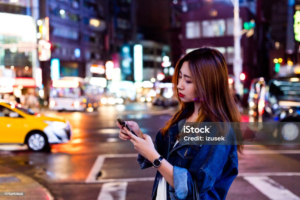 Woman using mobile phone while crossing street Side view of woman using mobile phone while crossing street at night. Beautiful female is text messaging on smart phone in city. She is wearing denim jacket. 20-24 Years Stock Photo