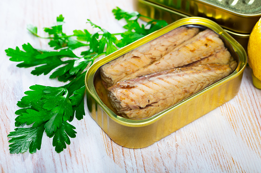 Appetizing scomber fillets in sunflower oil in open tin can on wooden table with fresh parsley and lemon