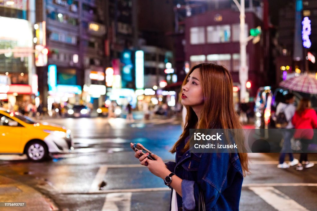 Young woman with mobile phone crossing city street Side view of beautiful woman holding mobile phone while looking away. Urban female is crossing city street at night. She is having long brown hair. Brown Hair Stock Photo