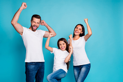 Portrait of cheerful family having brunette hair raising fists screaming, yeah celebrating victory wearing white t-shirt denim jeans isolated over blue background