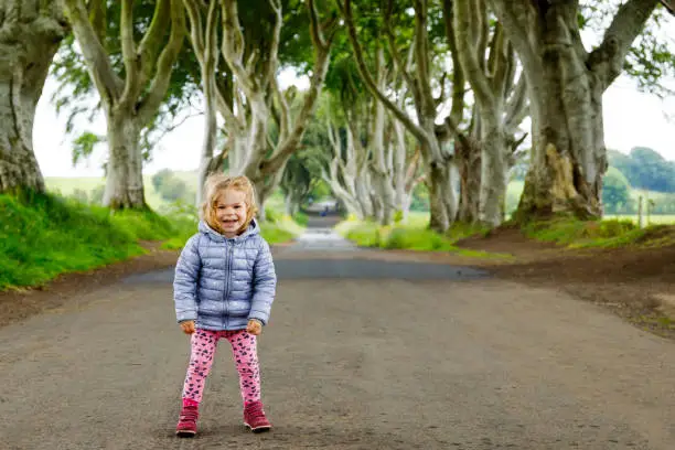 Photo of Cute toddler girl walking on a rainy day in the beginning of The Dark Hedges. Northern Ireland. Happy child visiting with parents and family famous Irish tree avenue
