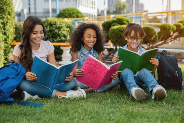 屋外の草の公園に座ってバックパックやノートを持つかわいい幸せな小さな学校の子供たち - multi ethnic group teenage girls little boys child ストックフォトと画像