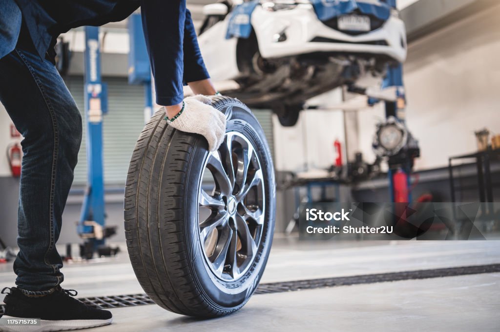 Male mechanic hold and rolling tire at repairing service garage background. Technician man replacing winter and summer tyre for safety road trip. Transportation and automotive maintenance concept Tire - Vehicle Part Stock Photo