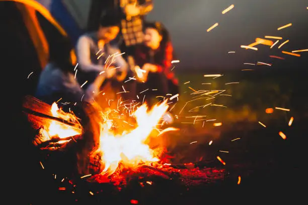 Photo of Sparking bonfire with tourist people sit around bright bonfire near camping tent in forest in summer night background. Group of student at outdoor fire fuel. Travel  activity and long vacation weekend