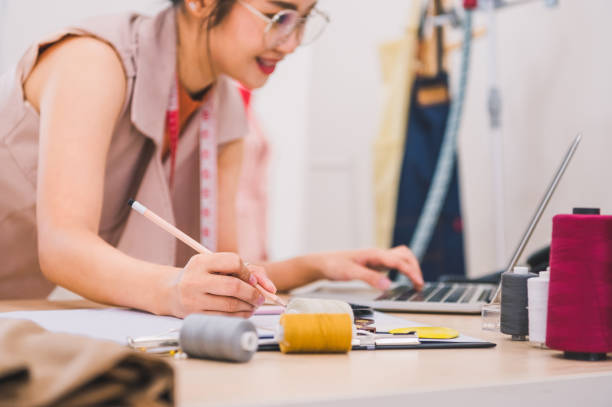 幸せなアジアの女性ファッションデザイナーは、衣料品店のスタジオで働き、顧客に連絡するためにラップトップコンピュータを使用しています。デザイン新しいコレクションを作成する女� - owner embroidery small business indoors ストックフォトと画像