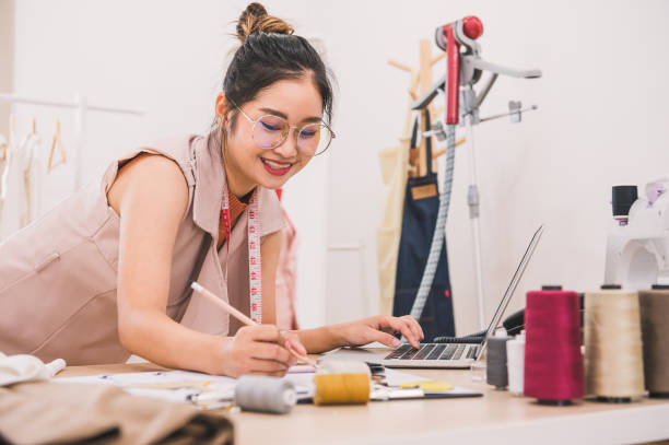 幸せなアジアの女性ファッションデザイナーは、衣料品店のスタジオで働き、顧客に連絡するためにラップトップコンピュータを使用しています。デザイン新しいコレクションを作成する女� - owner embroidery small business indoors ストックフォトと画像