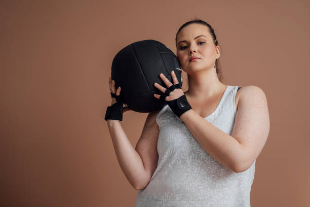 Plus Size Woman Posing with Medicine Ball Beautiful Caucasian plus size woman in sportswear holding fitness ball and looking at camera. body confidence stock pictures, royalty-free photos & images