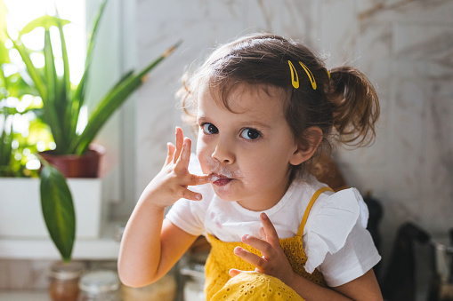 Portrait of cute Caucasian little girl eating whipped cream with her fingers and looking at camera.