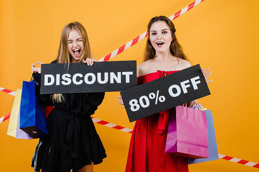 two screaming women have discount 80% off sign with colorful shopping bags and signal tape isolated over yellow