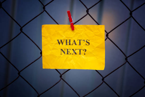 Paper note with question What is next hanging on a metal mesh fence. Close up.