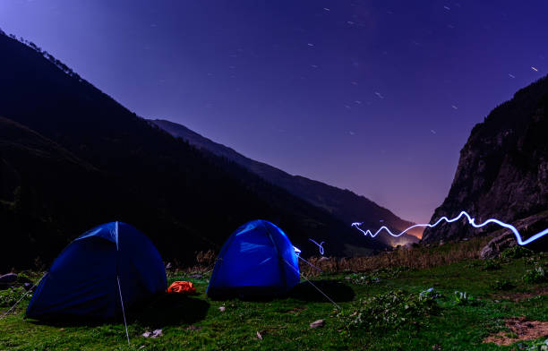 Night mountain landscape with illuminated blue tent &flash light trails. Silhouettes of mountain peaks and edges night sky with many stars & blue tent on foreground Night mountain landscape with illuminated blue tent &flash light trails. Silhouettes of mountain peaks and edges night sky with many stars & blue tent on foreground Glamping in Himachal stock pictures, royalty-free photos & images
