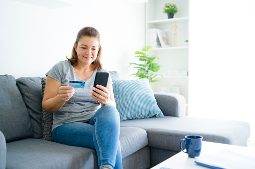 Online shopping: middle aged Hispanic woman sitting on couch holding credit card and mobile phone. The composition is at the left of an horizontal frame leaving useful copy space for text and/or logo at the right. Predominant color is blue. XXXL 42Mp studio photo taken with SONY A7rII and Zeiss Batis 40mm F2.0 CF