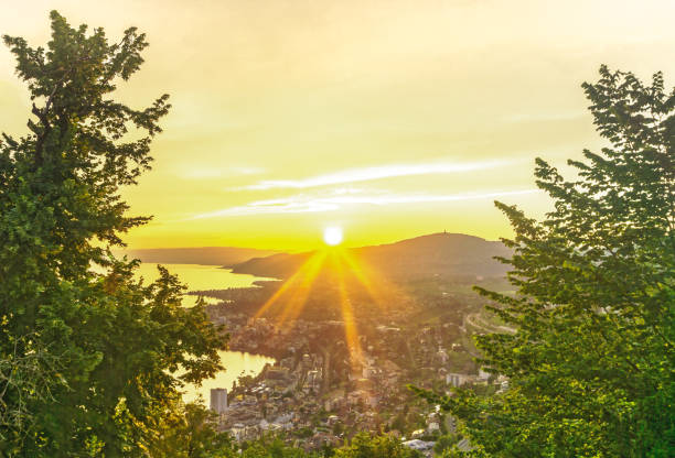 Bright yellow rays of the rising sun illuminate the city and the lake. View from the mountain through the leaves of trees