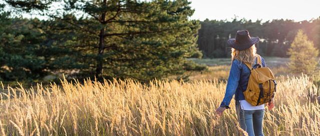 Woman wearing jeans, denim jacket, hat and backpack is standing in nature during sunset. Female tourist enjoying hiking