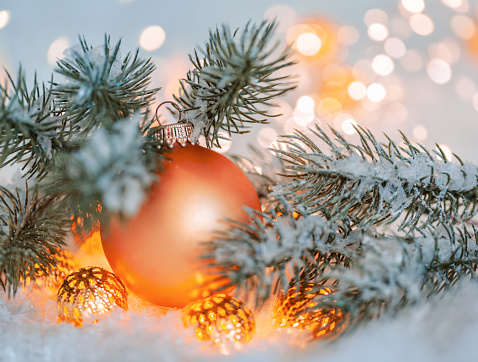 Holiday greeting card. Beautiful orange ball, pine branches and a garland in the snow. Blurred yellow bokeh background.