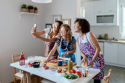 Group of Multiracial Female Friends Taking Selfie While Preparing Food at Home. Handsome Mixed Races Female Friends Making Selfie and Smiling While  Making Pizza in Kitchen Together. Multi-ethnic Female Friends Having Fun at Home