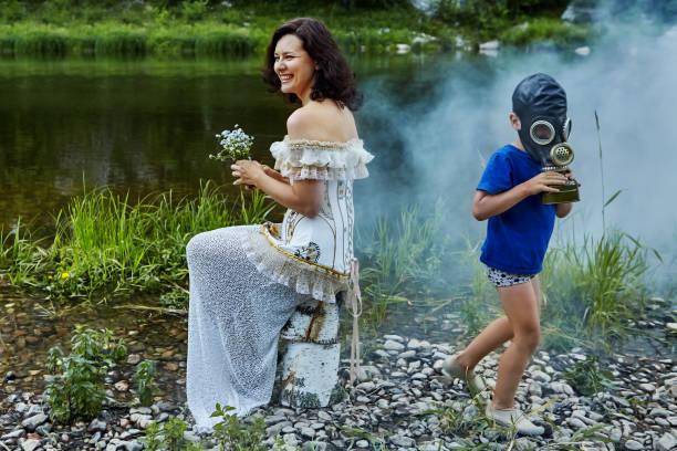 A beautiful adult woman laughs, sitting on the riverbank with a bouquet of wildflowers in her hands, wearing a wedding or evening dress, and a child in a protective gas mask is running nearby.