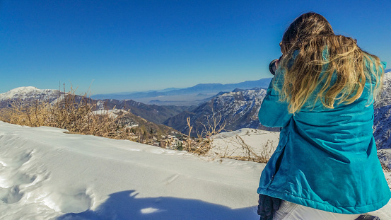 Rear view of a Brazilian Woman on her knees in ski clothes photographing a snowy landscape in Farellones/Chile.