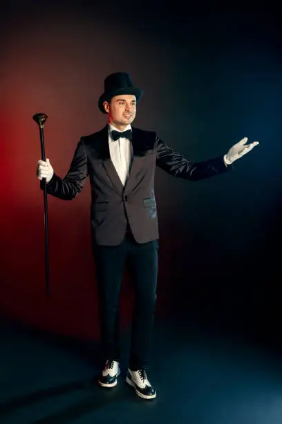 Professional showman wearing suit gloves and top hat standing isolated on black and red background holding cane hands aside greeting show begins smiling cheerful full body shot.
