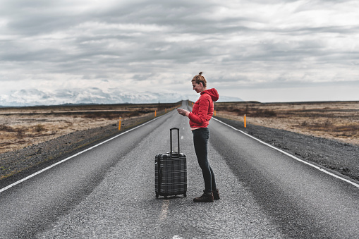 Woman in a red jacket with suitcase checking her phone on an empty road in a middle of nowhere.