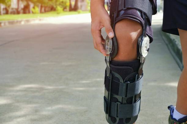 Man's leg with supportive knee brace on jogging in public park area Young man's hand turns angle movement control of supportive knee brace on his leg for jogging with flare light and blurred public park background, exercise and health care concept knee brace stock pictures, royalty-free photos & images