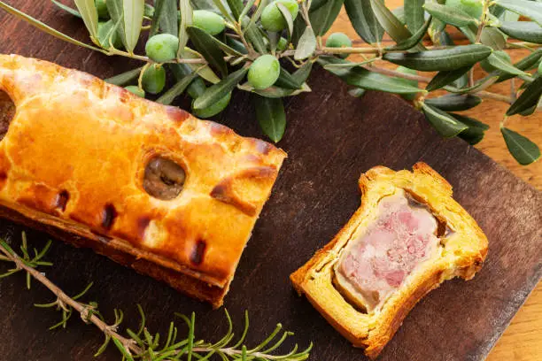 Top view of a pate en croute or pâté en croûte or meat pie, with rosemary twig and green olives on branch with leaves over a dark wooden cutting board and a used oak wood background. French traditional appetiser.