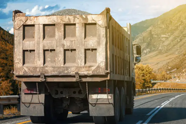 Rear view on a large dump truck rides on a highway in the mountains while transporting goods over long distances. Fast delivery by ground transportation.