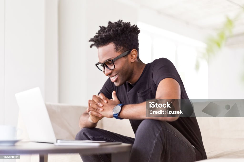 Businessman with hands clasped looking at laptop Smiling male entrepreneur using laptop while working at creative office. Confident afro hipster businessman is sitting on sofa at workplace. He is with hands clasped. 30-34 Years Stock Photo