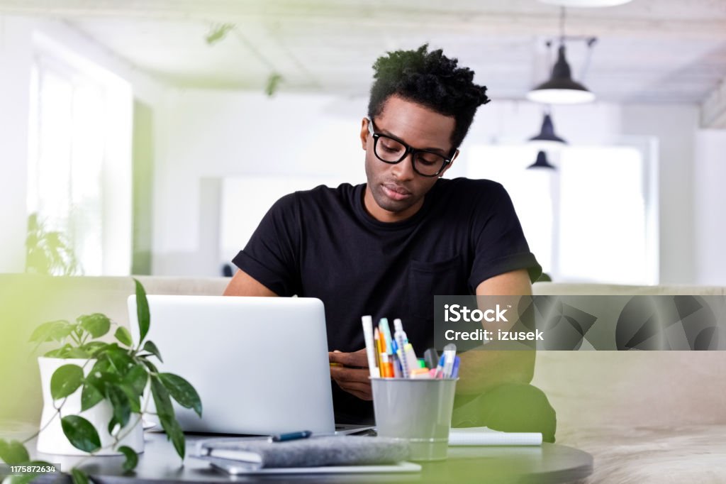 Confident businessman sitting with laptop on sofa Confident businessman sitting with laptop on sofa at creative office. Afro male hipster financial advisor is working at workplace. He is in smart casual. 30-34 Years Stock Photo