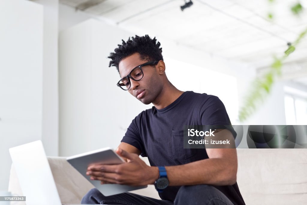 Male hipster professional using digital tablet Low angle view of confident hipster professional using digital tablet at workplace. Afro male executive is sitting on sofa at creative office. He is in smart casual. 30-34 Years Stock Photo