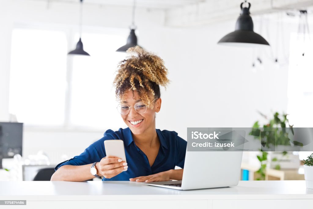 Smiling female hipster executive using smart phone Smiling female hipster entrepreneur using smart phone while sitting with laptop. Businesswoman is working at desk in creative office. She is with curly blond hair at workplace. Financial Advisor Stock Photo