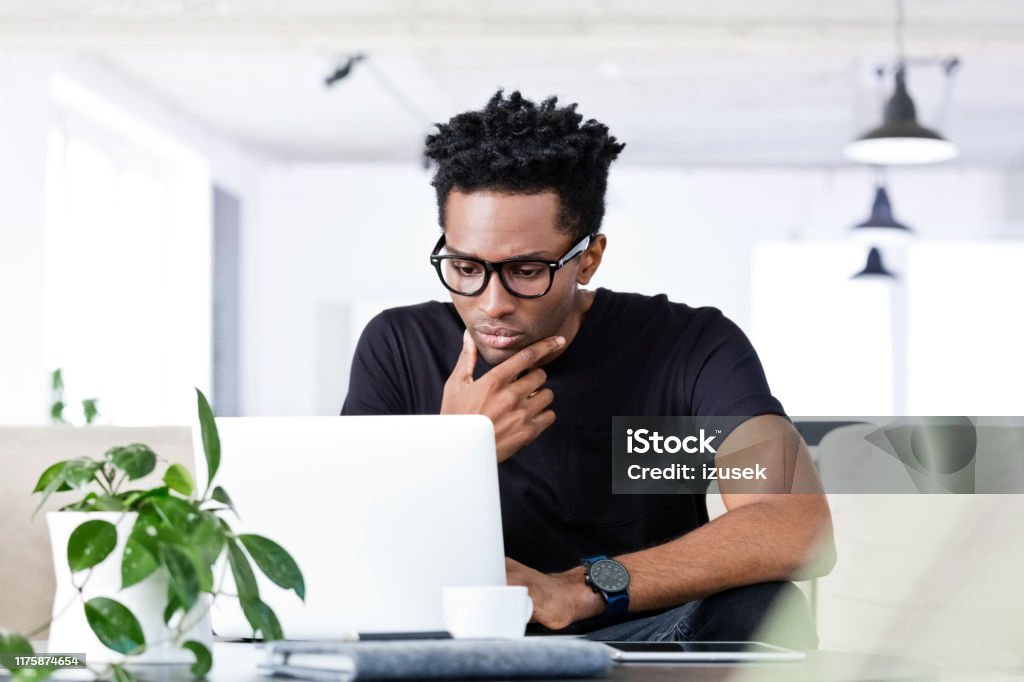 Thoughtful afro businessman using laptop Thoughtful male hipster executive using laptop at creative workplace. Afro businessman is sitting with hand on chin at office. He is in smart casual. 30-34 Years Stock Photo