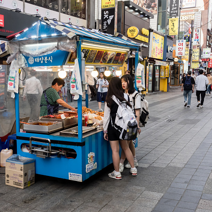 Suwon, South Korea - June 15, 2017: Vendor woman sells food to two young girls in her fast food kiosk at main street in Suwon. Street food is very popular in Korea.